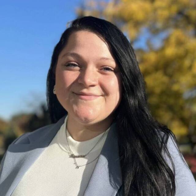 A woman with long black hair, silver necklaces, a white turtleneck, and a light blue jacket smiling faintly in front of a tree with fall leaves